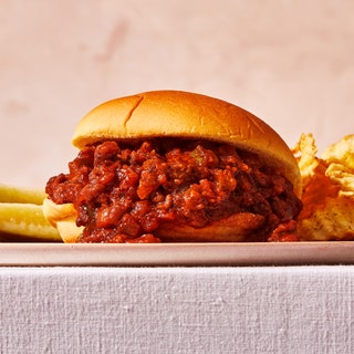 Closeup of a gochujang sloppy joe on a bun with ridged potato chips and pickles behind it