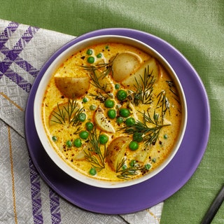 Bowl of potato chowder with peas garnished with dill sitting on top of a purple plate with a spoon off to the side.